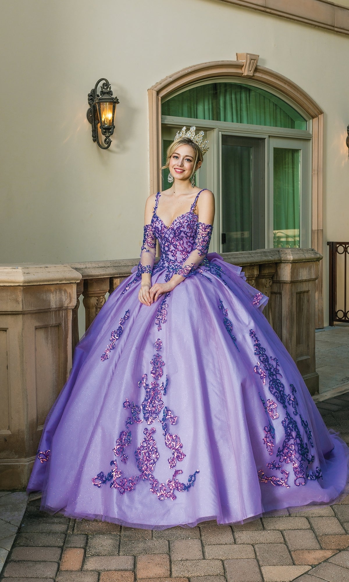 A young woman smiles in front of a building wearing the Quinceanera Dress 1652 By Dancing Queen, a jeweled tiara, and an ornate purple ball gown with embroidery and a sweetheart neckline.